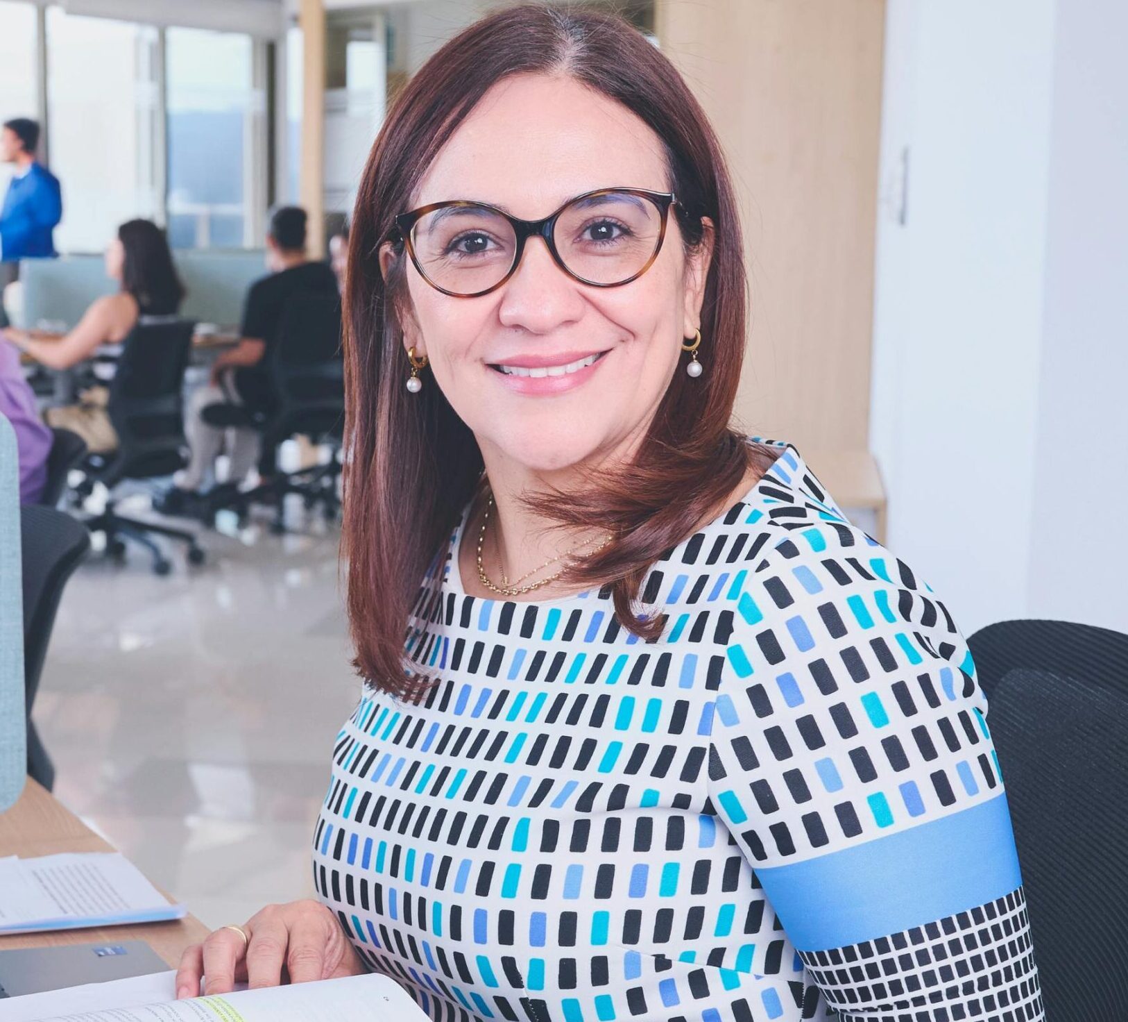 Confident woman in a modern office, surrounded by colleagues, working on a laptop.